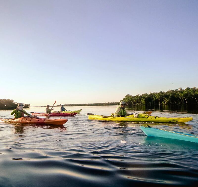 The image shows three people kayaking on a calm body of water. The kayakers are spread out, with one in the foreground and two further back. The water is reflecting the sky, and there are trees visible in the background. The sky is clear and blue, suggesting a sunny day. The kayaks are different colors, including red, yellow, and blue.